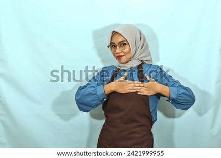 young Asian Muslim woman in hijab , glasses and brown apron, standing holding hands folded on heart and looking camera isolated on white background. muslim housewife concept