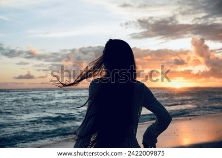 Back view of a woman gazing at the sunset over the ocean, her hair tousled by the sea breeze