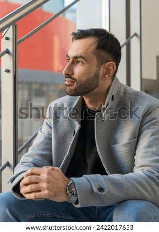 Portrait of a handsome young man. Smiling Asian man with a neat beard sits on the steps near the window and looks into the distance
