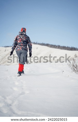 A woman in a colorful jacket and red leggings walks through a snowy field toward a hill on a bright winter day, enjoying the peaceful and serene landscape and embracing the tranquility of nature.