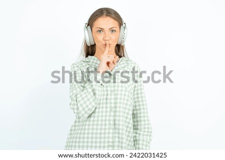 Young beautiful woman wearing green plaid pyjama making hush gesture with finger on her lips wearing  wireless headphones. Be quiet. Royalty-Free Stock Photo #2422031425