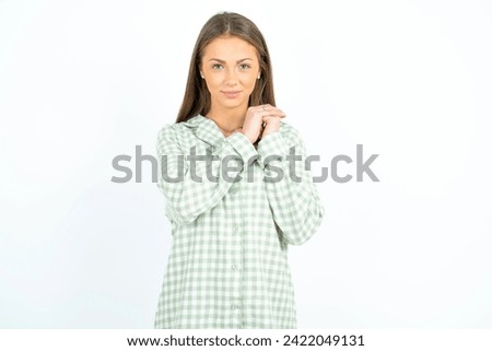 Charming serious Young beautiful woman wearing green plaid pyjama keeps hands near face smiles tenderly at camera