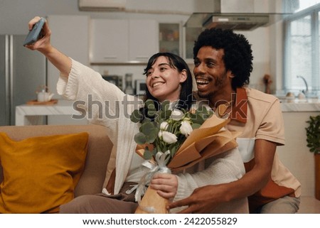 Young smiling woman with white flowers taking selfie with her husband at home while holding smartphone in front of herself