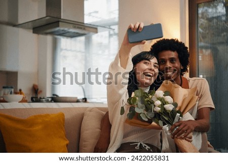 Happy wife with bouquet of flowers holding smartphone in front of herself and taking selfie with affectionate husband sitting next to her