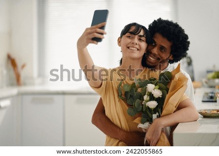 Young cheerful brunette woman holding smartphone while taking selfie by kitchen table with affectionate husband embracing her