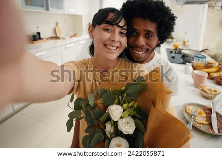 Young smiling man embracing his pretty wife with bunch of white flowers and both looking at camera while woman taking selfie