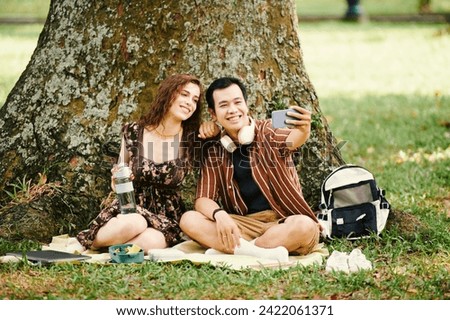Happy young affectionate couple looking at screen of smartphone held by Asian guy taking selfie with his girlfriend under tree in park