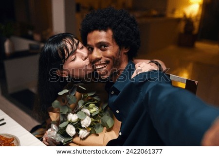 Young smiling African American man looking at camera while taking selfie with his affectionate wife with flowers kissing him on cheek