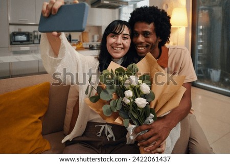 Happy young woman with bouquet of white flowers holding smartphone in front of herself while taking selfie with affectionate husband