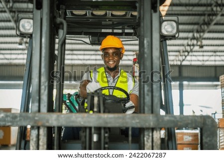 African forklift driver focused on carefully transporting stock from shelves of a large warehouse wearing a helmet and vest looking toward goods