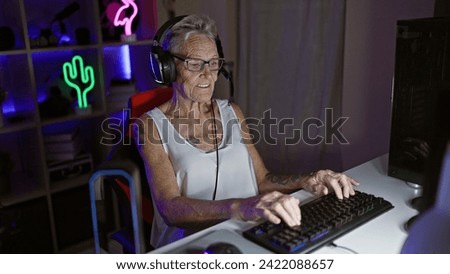 Confident, grey-haired senior woman streamer smiling while playing futuristic video game at night in gaming room, sitting indoors entertaining with modern technology