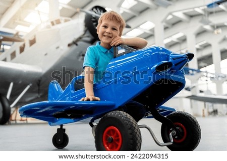 Portrait of cute little blond happy kid boy enjoy have fun play riding pedal toy plane model against vintage airplane background in museum hangar. Future child profession dream concept Royalty-Free Stock Photo #2422094315