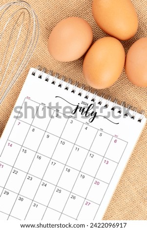 July 2024 Desk calendar and fresh eggs with egg whisk on burlap background.