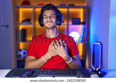 Young hispanic man playing video games smiling with hands on chest with closed eyes and grateful gesture on face. health concept. 