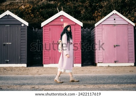 Stylish hipster woman with color hair in pink outfit and backpack walking along wooden beach huts on seaside. Off season Travel concept. Seasonal street fashion. Barbiecore style. Simple pleasures Royalty-Free Stock Photo #2422117619