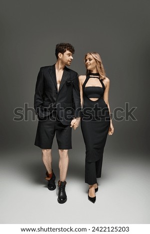 full length of couple in trendy black attire holding hands while standing on dark grey background