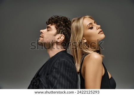 pretty woman sitting back to back with curly man in pinstripe blazer on grey backdrop, couple