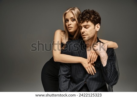 beautiful couple in elegant attire, woman in black dress embracing handsome man on grey backdrop