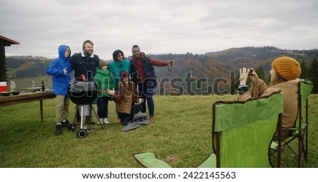 Multiethnic group of travelers stand near barbecue grill posing on camera. Woman in camp chair takes picture on phone. Big family resting outside of holiday house in the mountains. Tourism and travel.