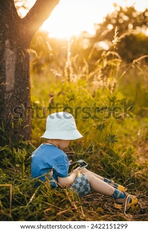 a little boy sits on the grass and holds a phone in his hands