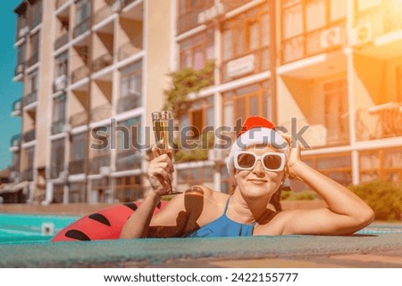 Woman pool Santa hat. A happy woman in a blue bikini, a red and white Santa hat and sunglasses poses near the pool with a glass of champagne standing nearby. Christmas holidays concept.