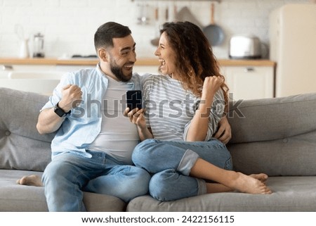 Overjoyed young couple sit on couch in kitchen triumph read good unexpected news on cellphone gadget, excited millennial husband and wife feel euphoric win online lottery on smartphone, luck concept Royalty-Free Stock Photo #2422156115