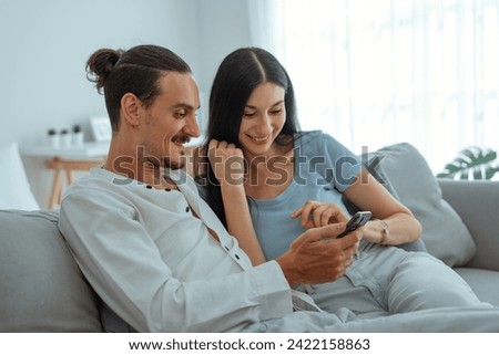 Couple enjoying watching online content in a smart phone sitting on sofa at home in the living room, positive man holding smartphone showing funny online photo to girlfriend spending weekend together