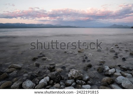 Long exposure shot of Nahuel Huapi lake at sunset. Beautiful blurred water effect, the rocky shore and Andes mountains in the horizon with a magical dusk light.