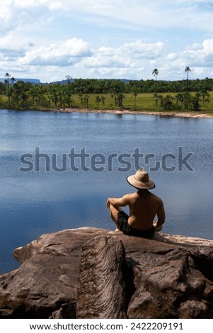 adult male backwards shirtless with hat sitting on edge of rock in Salto El Sapo waterfall with beautiful view of Canaima lagoon, Carrao river, Canaima National Park, Venezuela Royalty-Free Stock Photo #2422209191