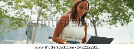 Cute tanned woman with long brown hair in white top and yellow bandana using laptop while sitting on bench. Pretty girl student preparing for classes in outside, Panorama
