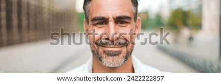 Close-up mature businessman with neat beard is looking at the camera in the financial district in the city, Panorama. Successful man smiling at the camera on modern buildings background