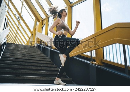 Vivacious young Caucasian woman energetically ascending stairs in bright, geometric yellow overpass, fitness-focused with water bottle in hand, capturing motion and health Royalty-Free Stock Photo #2422237129