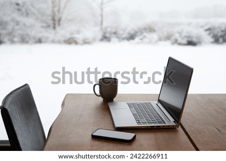 Laptop, smart phone and cup of coffee on wooden table. Concept of working from home and remote work with winter landscape with snow outside. Photo taken in Sweden.