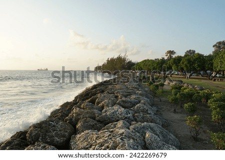 View on artificial breakwater built by rocks and piled up in wave breaker to protect Port of Bridgetown, Barbados from waves of Caribbean Sea. There are also some people and ship on horizon. Royalty-Free Stock Photo #2422269679