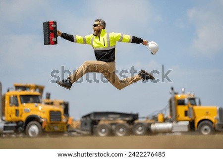Hispanic builder excited jump and run on site construction. Excited builder construction worker in a safety helmet jumping in front of the trucks. Excited crazy builder man in helmet jump outdoor. Royalty-Free Stock Photo #2422276485