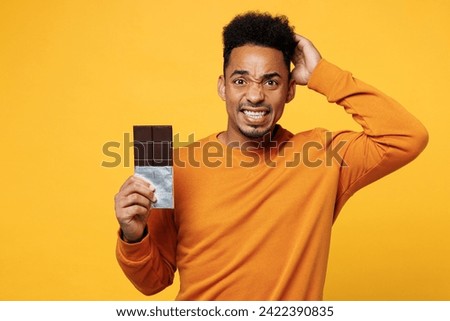 Young sad man he wear orange sweatshirt casual clothes hold eat bar of chocolate scratch head isolated on plain yellow background studio. Proper nutrition healthy fast food unhealthy choice concept