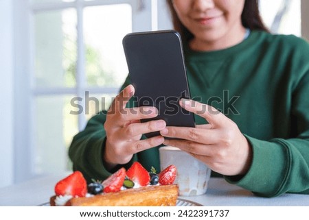 Closeup image of a young woman holding and using mobile phone with coffee cup and cake on the table