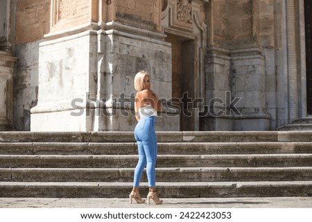 Pretty blonde adult woman dressed in jeans and white top having fun posing for photos on the steps of the cathedral in Cadiz, Spain. The woman is on a holiday trip