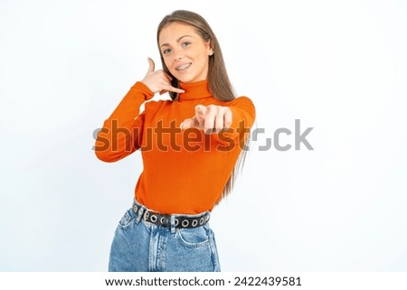 Young beautiful woman smiling cheerfully and pointing to camera while making a call you later gesture, talking on phone