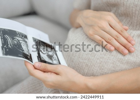 Close-up of a pregnant woman sitting in the living room at home looking at the ultrasound image of her own baby. Happy pregnancy woman during prenatal ultrasound at her home Royalty-Free Stock Photo #2422449911
