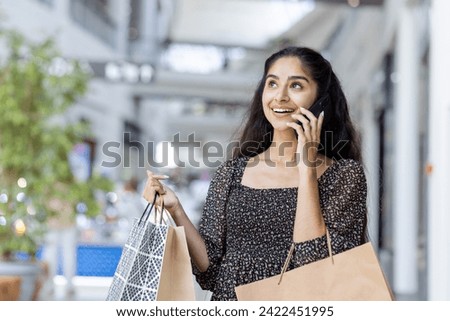 Smiling young Indian woman enjoying shopping, walking in a mall with colorful bags, chatting happily on her smartphone. Royalty-Free Stock Photo #2422451995