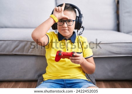Young hispanic kid playing video game holding controller wearing headphones making fun of people with fingers on forehead doing loser gesture mocking and insulting. 