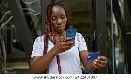 Casual yet serious, a beautiful african american woman with braids makes a note of her finances. in the urban street sunlight, she uses her smartphone to photograph her credit card.