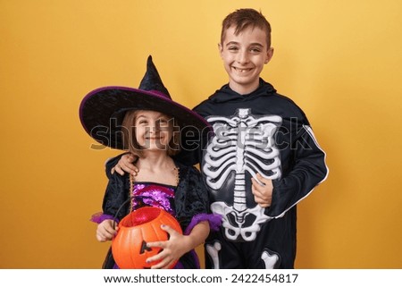 Delighted brother and sister wearing halloween costumes, sharing toothy, confident smiles over an isolated yellow background. embodying joy, happiness, and a positive outlook together. Royalty-Free Stock Photo #2422454817
