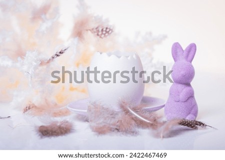 A small purple Easter bunny standing next to an open egg, candles standing on a purple plate, surrounded by feathers and boho style decorations on a light background and a white table