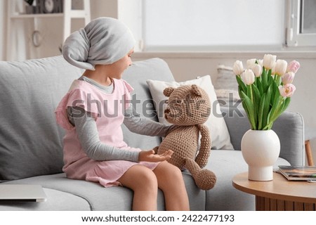 Little girl after chemotherapy with toy bear sitting on sofa at home. International Childhood Cancer Day Royalty-Free Stock Photo #2422477193