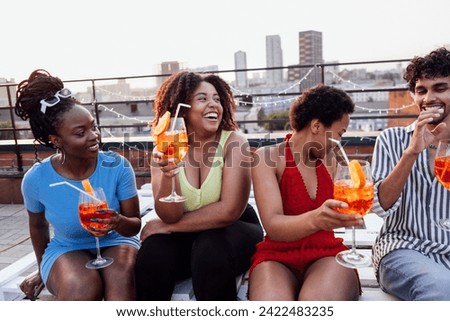 Company of four cheerful friends are enjoying tasty cocktails. Roof party. Interracial man and african women drink aperol with orange slices. Multiethnic group of people have fun and laugh together. Royalty-Free Stock Photo #2422483235