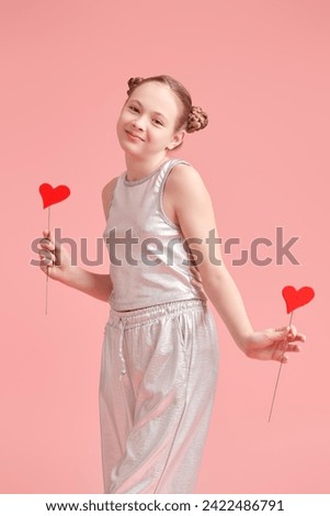 Cute teenage girl posing emotionally with red hearts, covering her eyes, on a pink studio background. Valentine's Day concept.