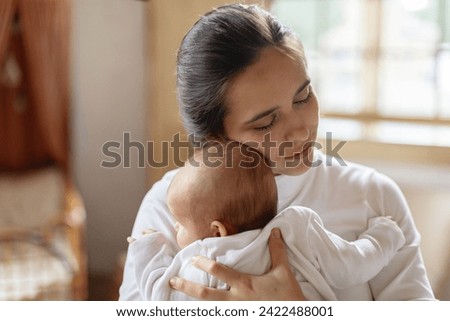 Pretty young woman holding her little baby daughter in her arms, lullying and embracing, bonding at home in bedroom interior