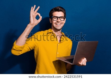 Photo portrait of nice young male netbook show okey symbol specs wear trendy yellow garment isolated on dark blue color background